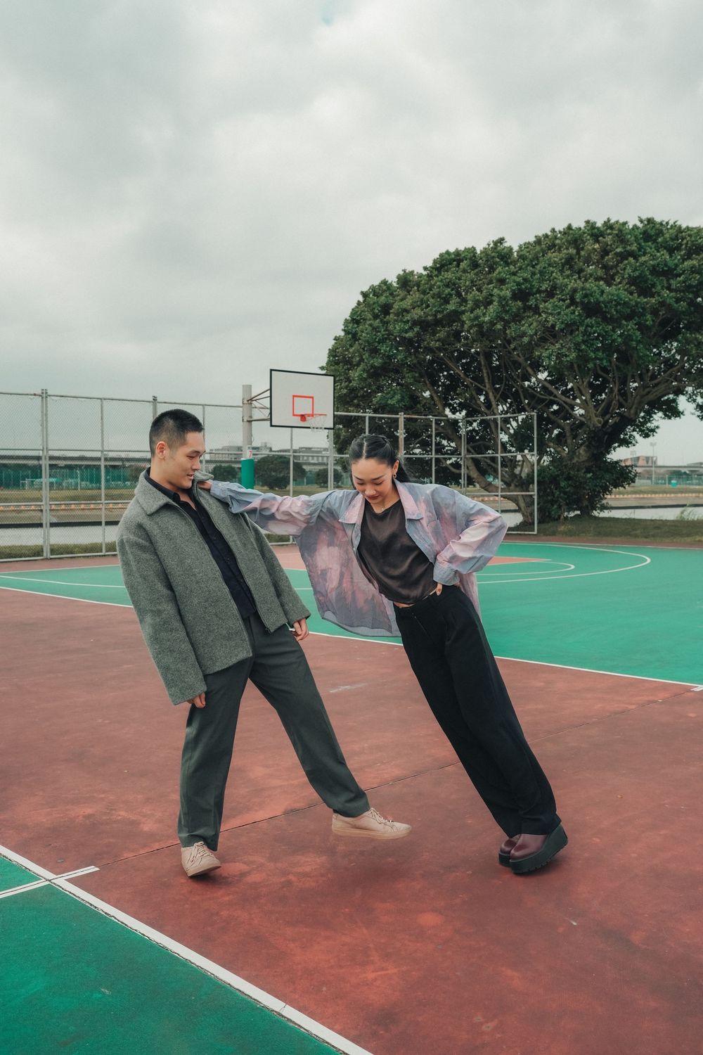 Two people in stylish casual wear perform playful dance moves on a green and red outdoor basketball court.
