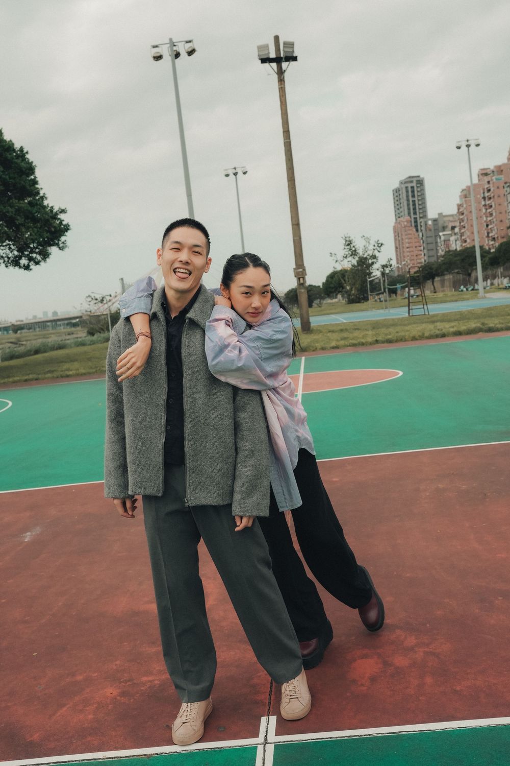 Two people in casual winter outfits playfully pose together on a green and red outdoor basketball court.