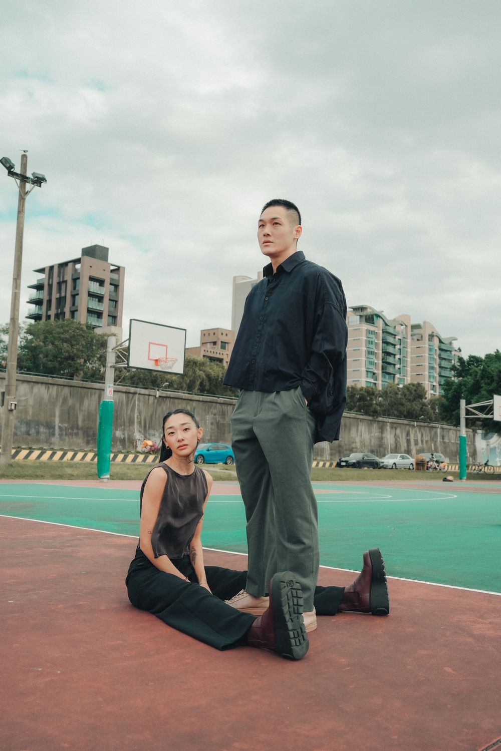 Two people pose on an outdoor basketball court with apartment buildings in the background on an overcast day.