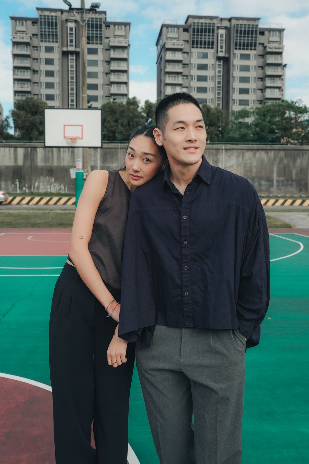 Pair of people in dark clothing posing together on an outdoor basketball court.