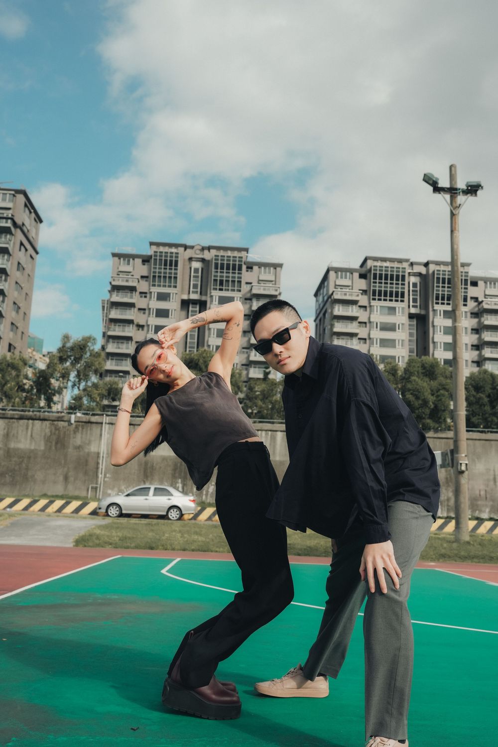Two people in black outfits strike dynamic poses on a green basketball court with urban buildings behind them.