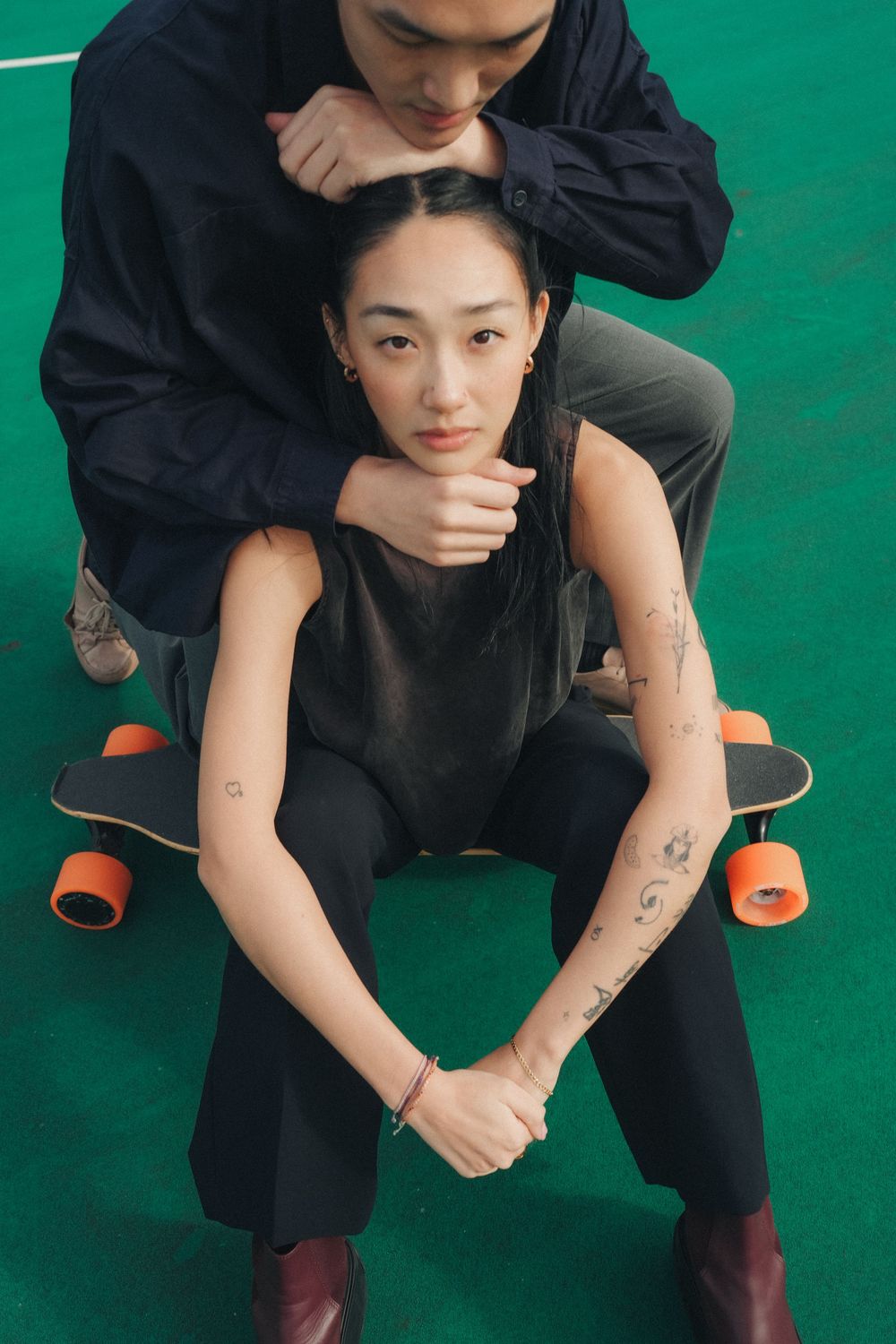 Stylish couple in dark clothing posing with a skateboard on green surface, showcasing arm tattoos and casual urban style.