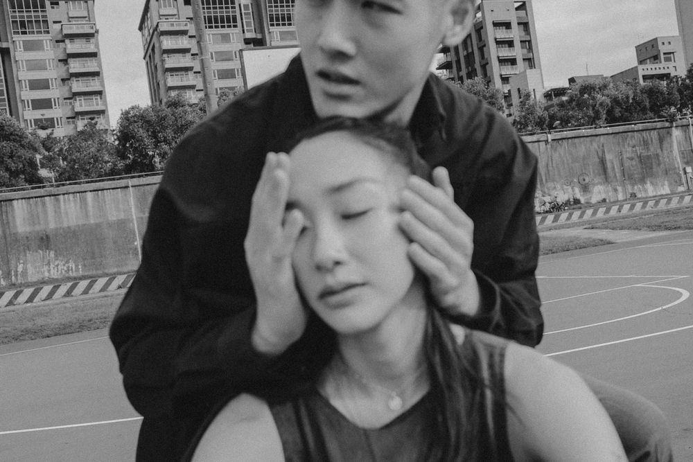 Two people share an intimate moment on a basketball court with urban apartment buildings in the background.