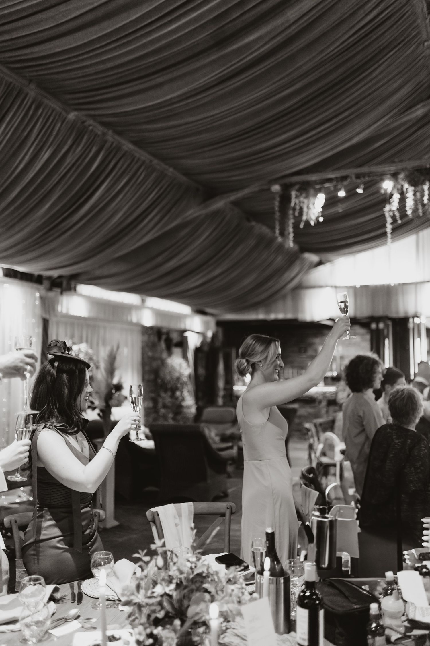 A black and white photograph of guests at a wedding reception with draped fabric ceiling and elegant table settings.
