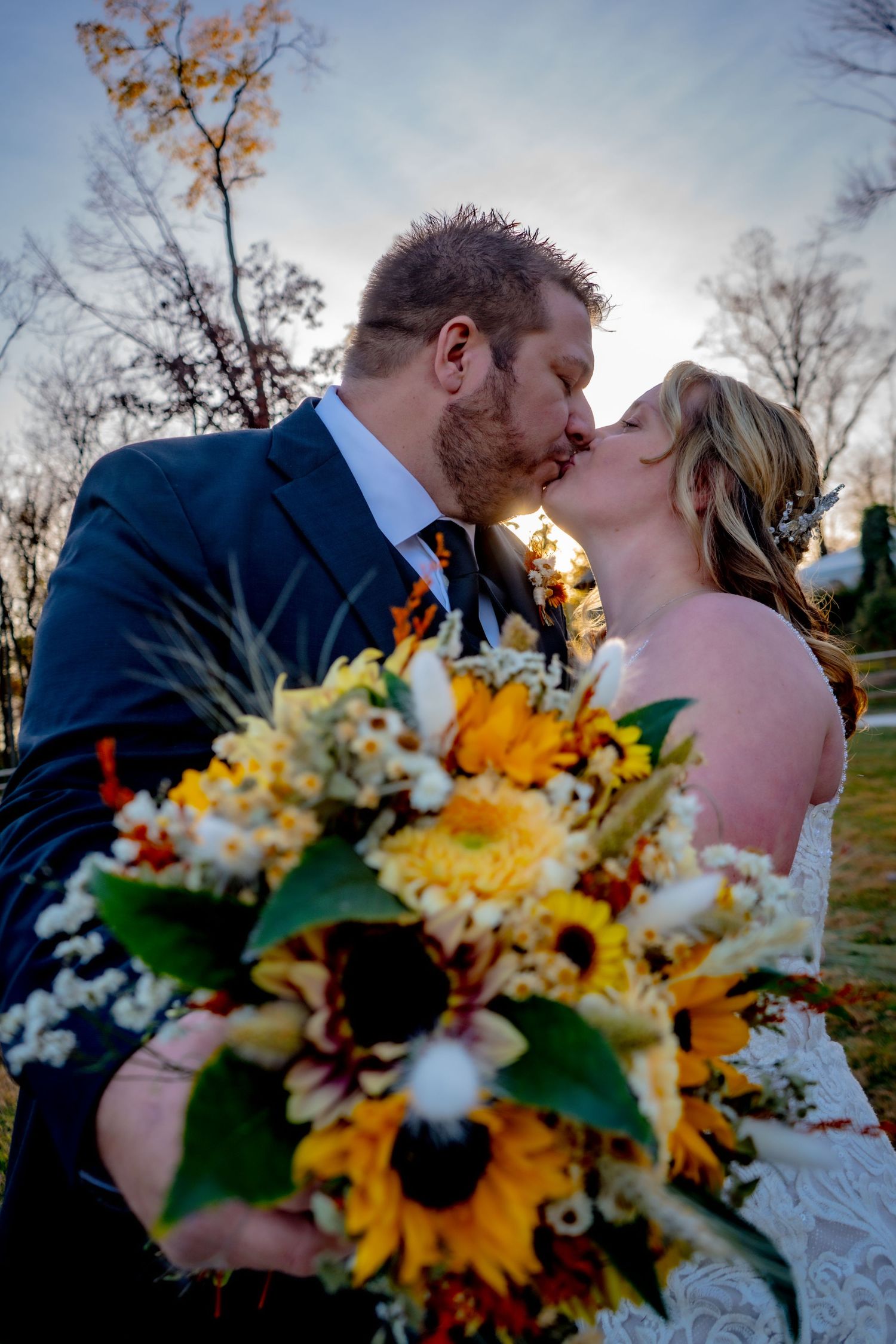 A couple in wedding attire stands silhouetted against a vibrant orange sunset behind a tree line.