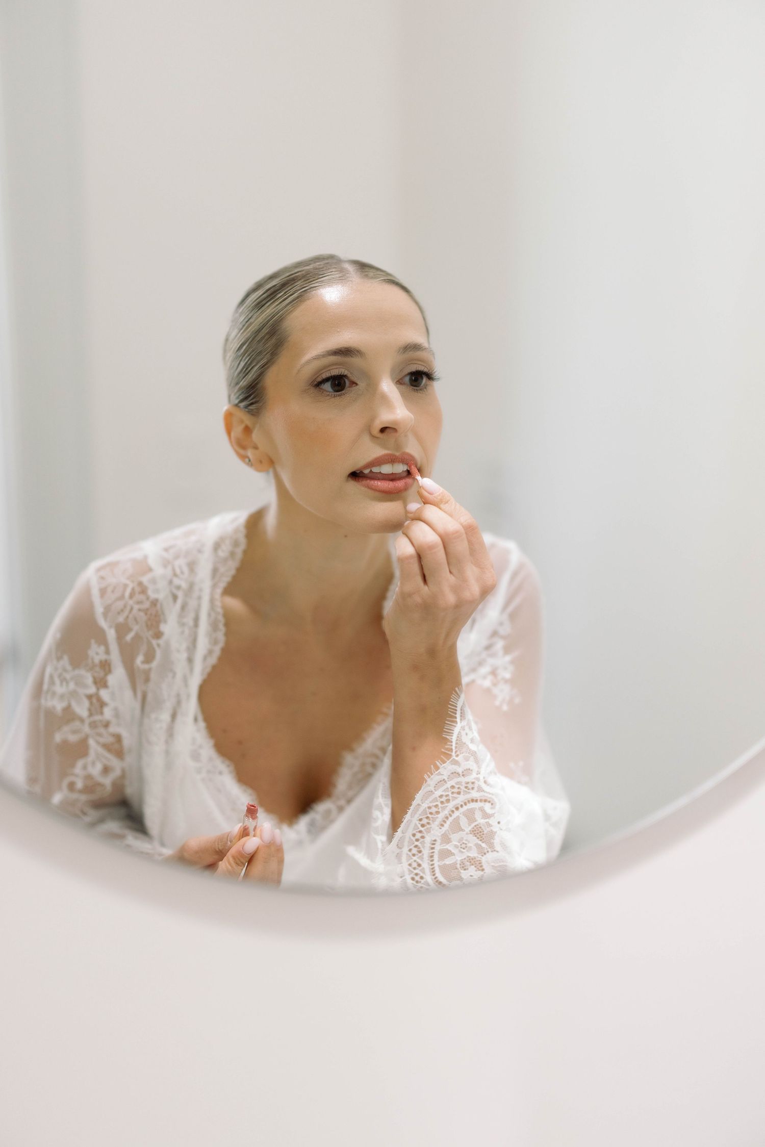 Woman in white lace robe applies makeup while looking in mirror during bridal preparation.