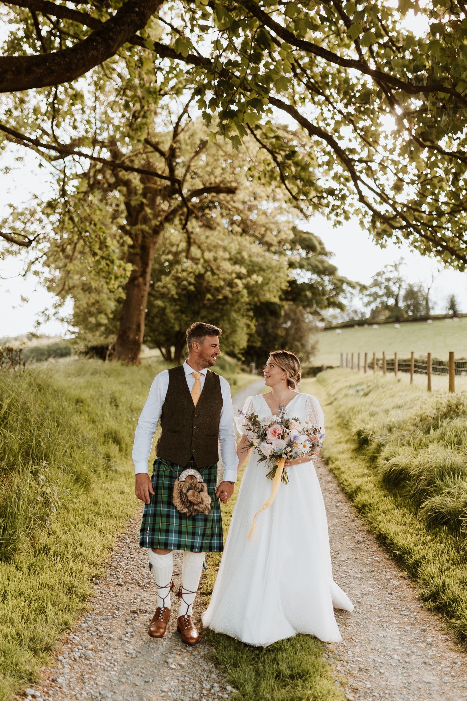 Two newlyweds walk down a tree-lined country path in traditional Scottish wedding attire with the groom wearing a tartan kilt.