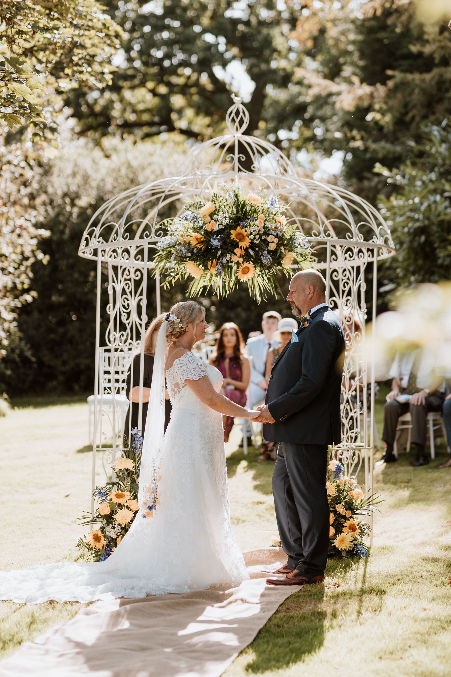 Beautiful outdoor garden wedding ceremony under an ornate white gazebo decorated with yellow and orange floral arrangements.