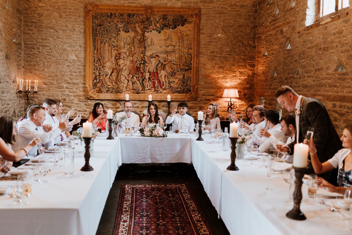A rustic Italian dinner party setup with long tables in a stone-walled room illuminated by candelabras and natural light.