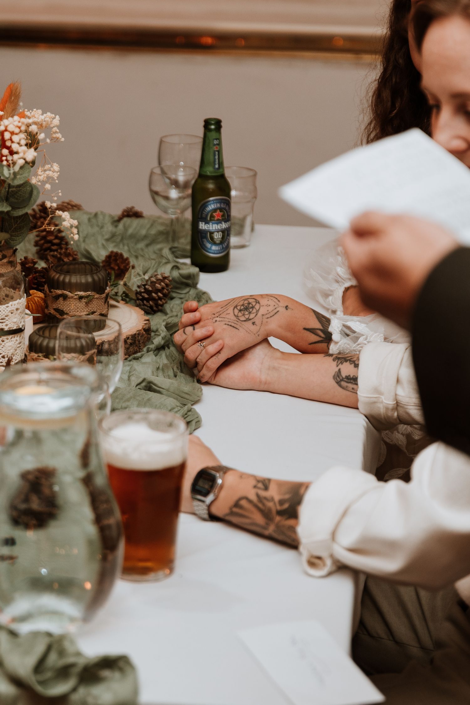 Moody dinner table setting with beer glasses, green foliage, and tattooed arms holding papers at a festive gathering.