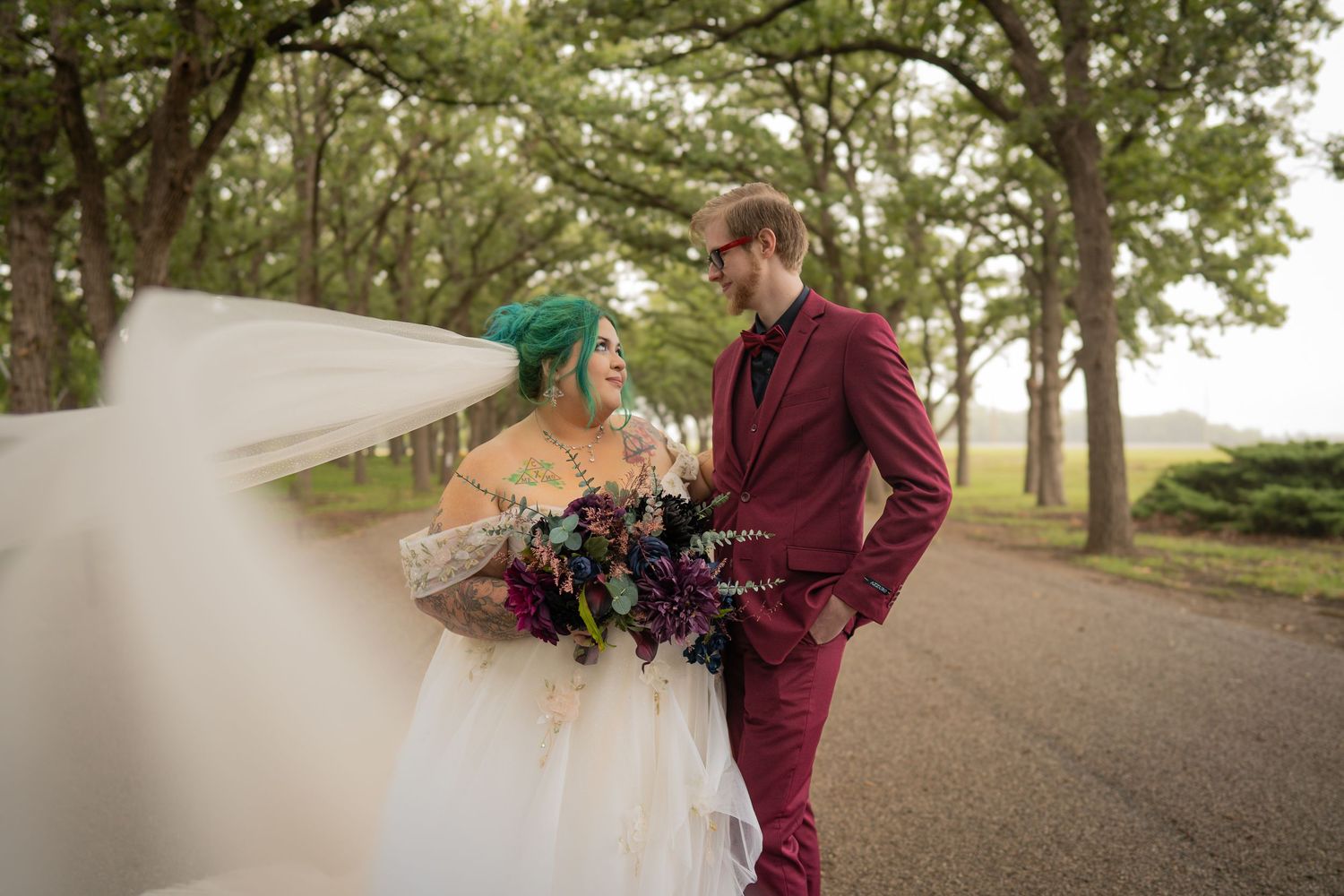 Wedding couple on tree-lined path with flowing veil in romantic pose.