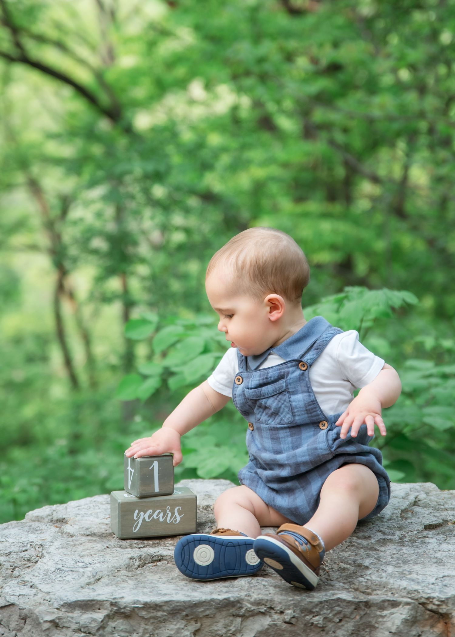 Toddler in denim overalls sits on a large rock exploring small decorative jar in lush green forest setting.