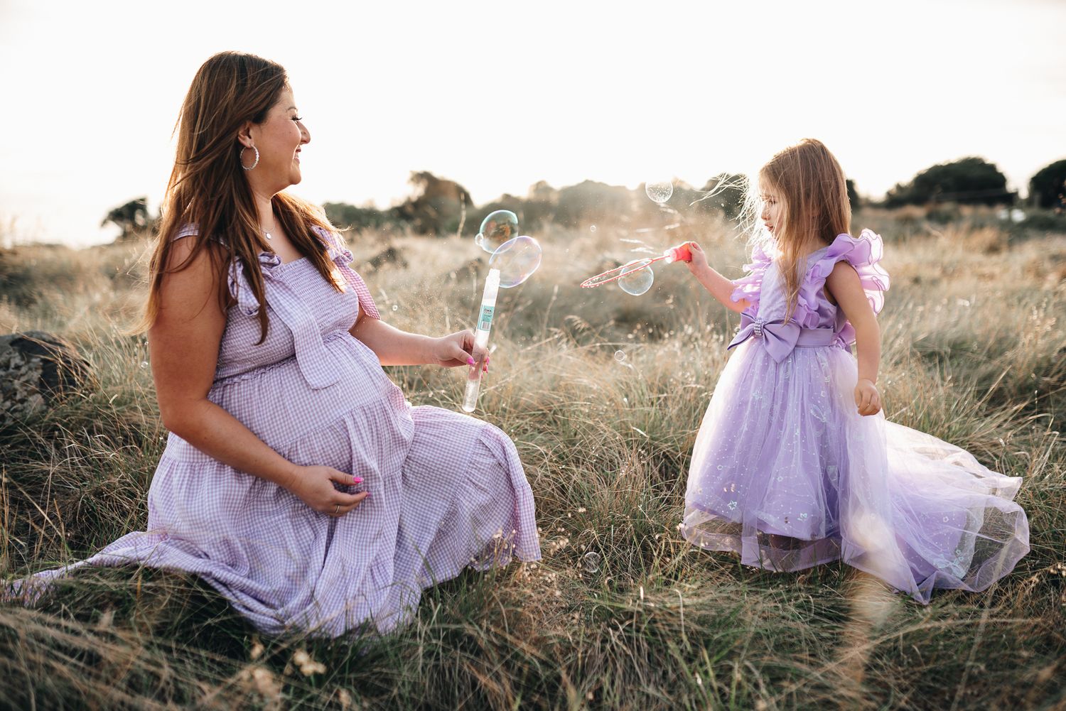 A pregnant woman in a flowing lavender dress in a sun-drenched meadow at golden hour taken by Melbourne maternity photographer.