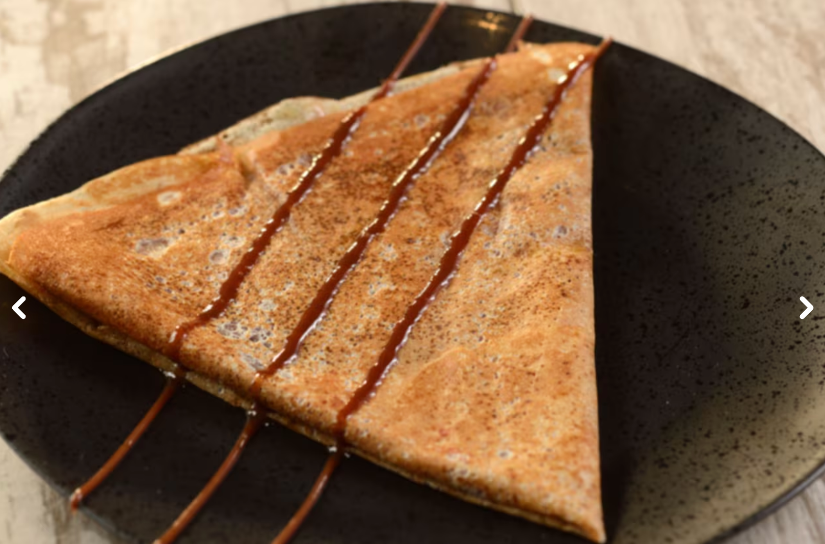 Close-up of chocolate-drizzled crepe on black plate with decorative pattern.