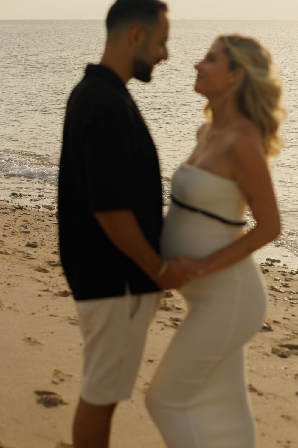 A couple shares an intimate moment on the beach at sunset, with the woman wearing a white dress and man in dark shirt.