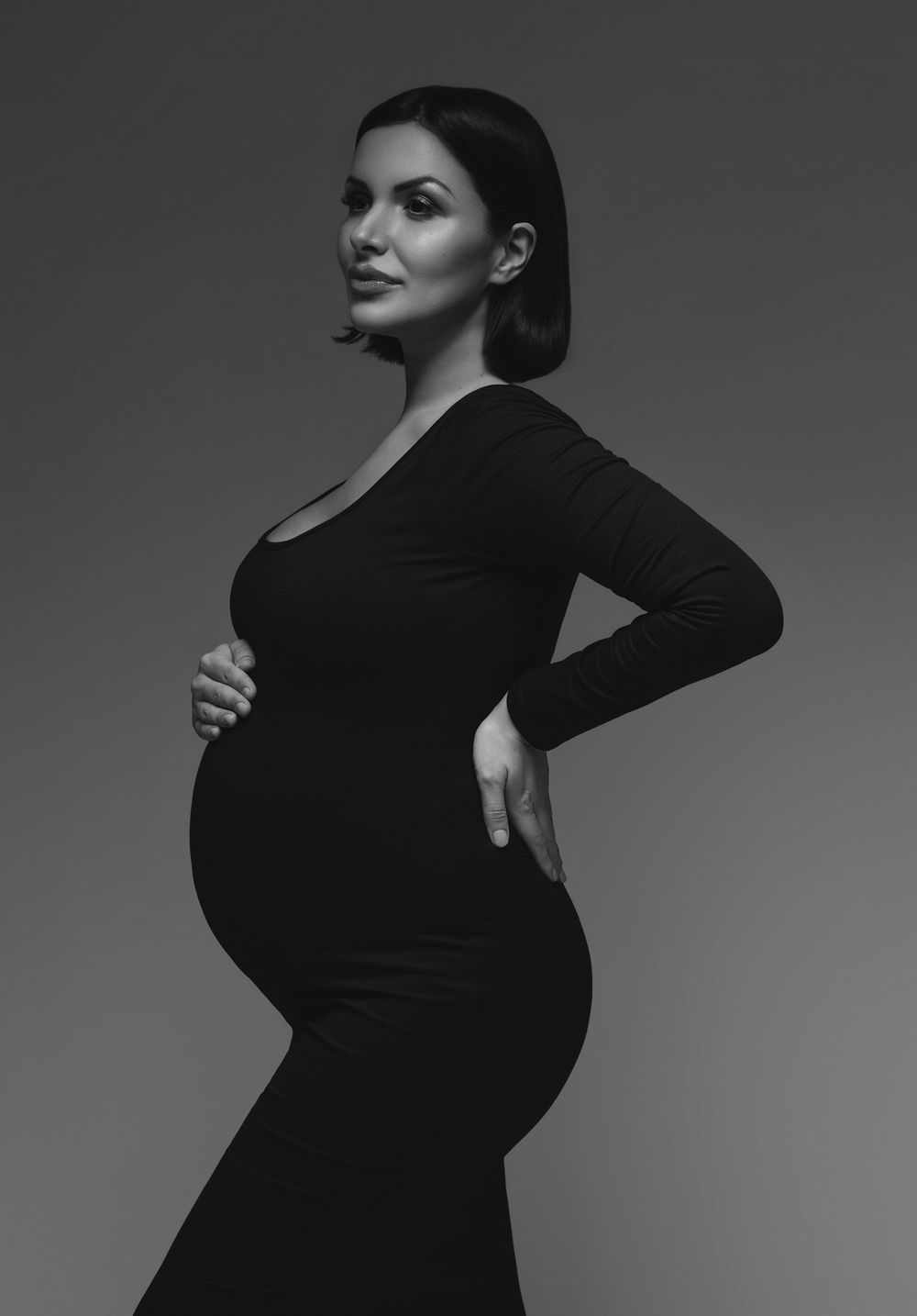 Black and white maternity portrait of a pregnant woman in a black dress cradling her baby bump.