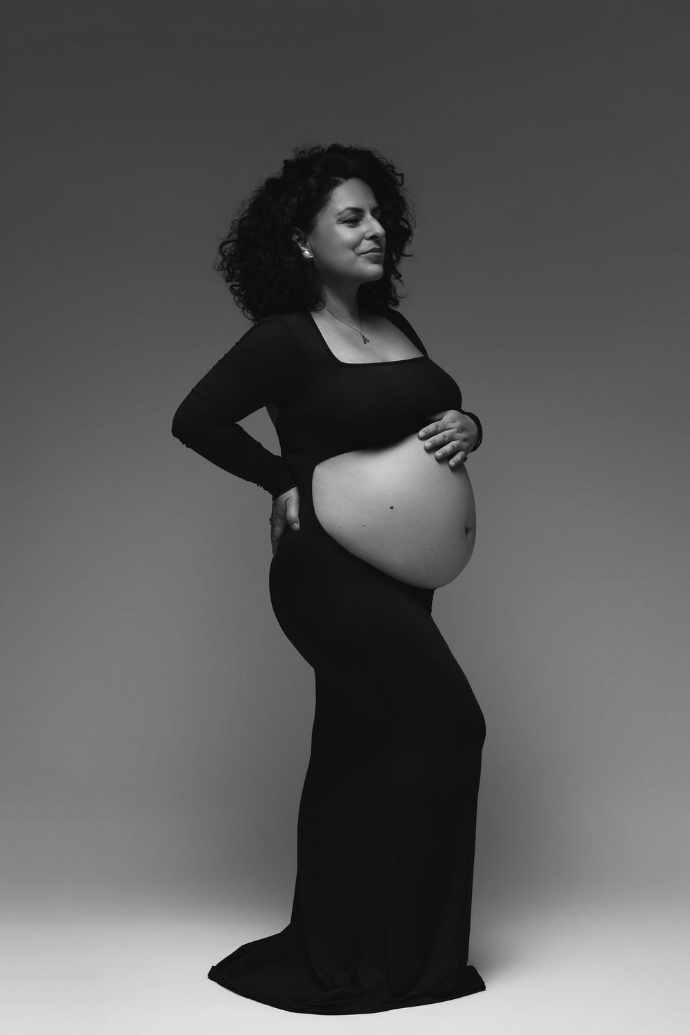 Pregnant woman in black gown cradling baby bump, elegant maternity portrait in black and white studio photography.