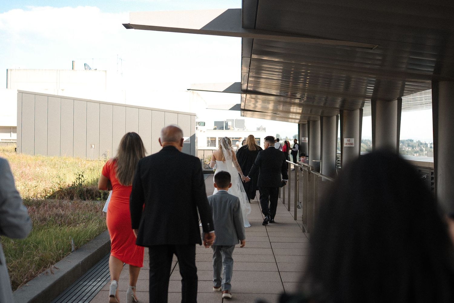 People walking along a covered outdoor walkway or breezeway along a modern building on a sunny day.