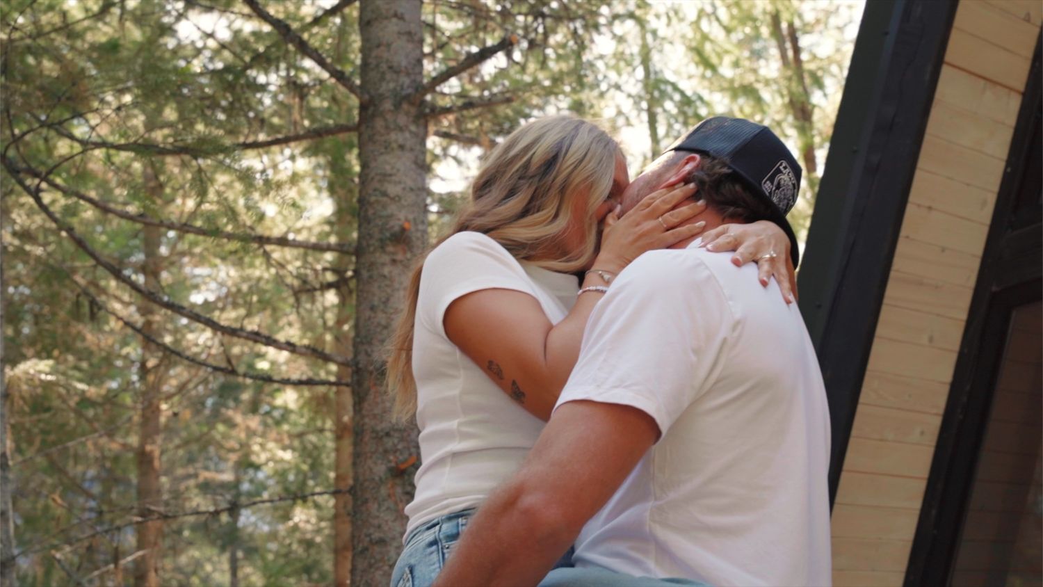 A romantic moment shared between a couple embracing on a wooden deck with tall pine trees in the background.