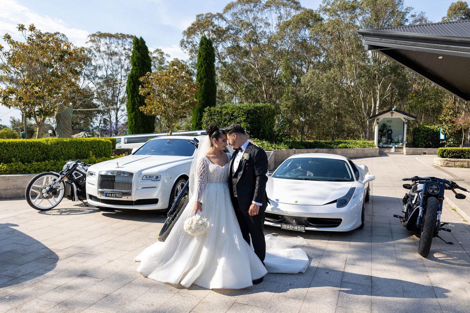 Wedding couple posing with luxury vehicles including Rolls-Royce and Ferrari at outdoor venue.