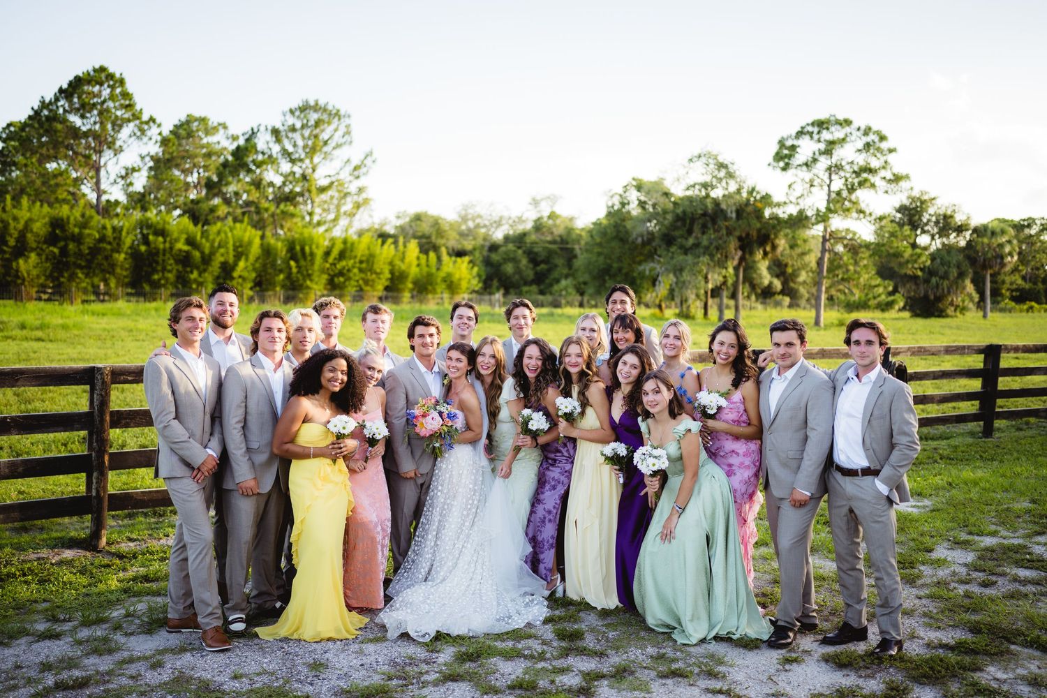 Large wedding party in pastel dresses and gray suits poses together on a sunny day by wooden fence at outdoor venue.
