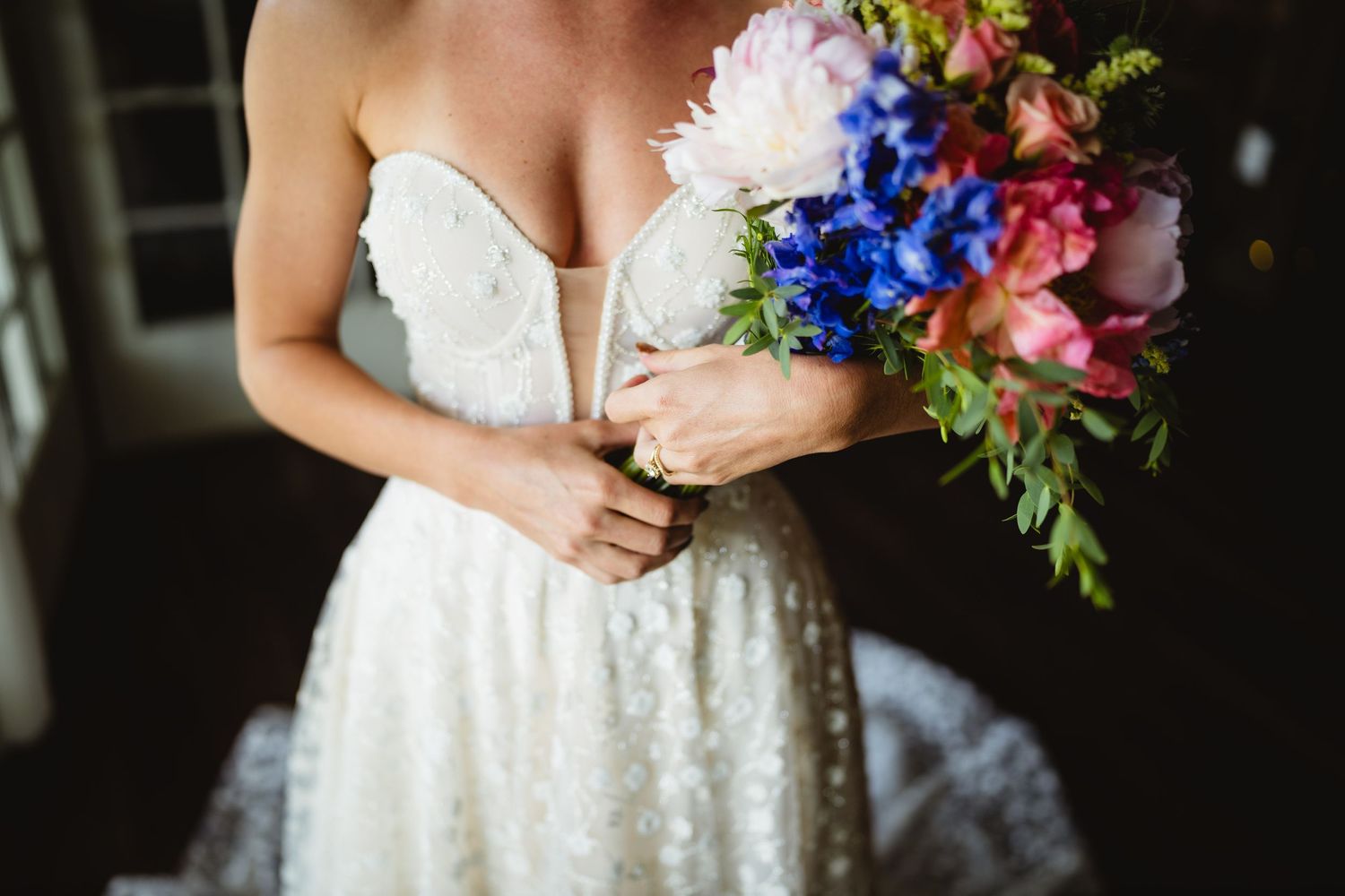 A bride holds a vibrant bouquet of pink, blue and white flowers paired with greenery against a white lace dress.