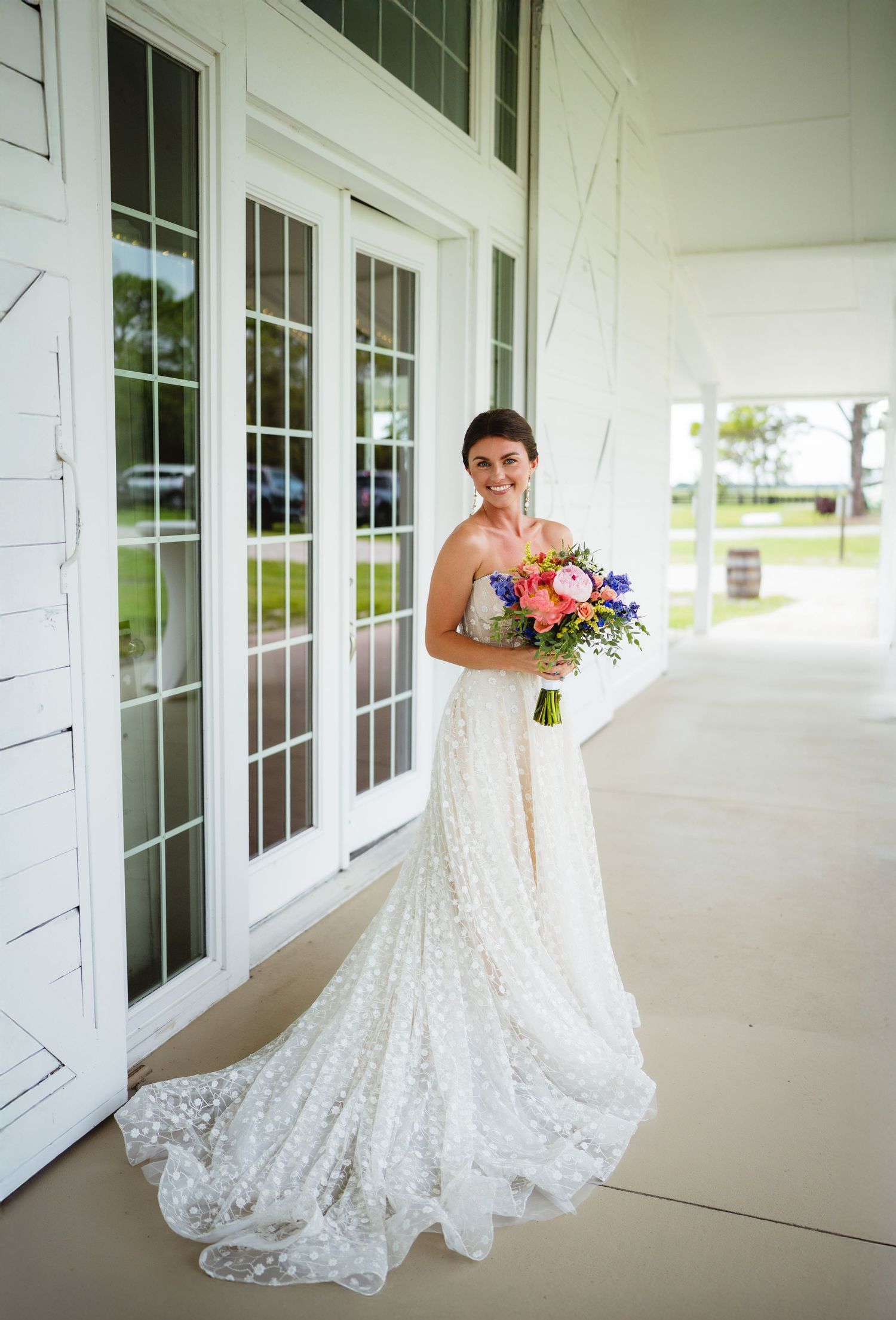 Bride in elegant lace wedding gown with long train poses on white porch holding colorful bouquet of summer flowers.