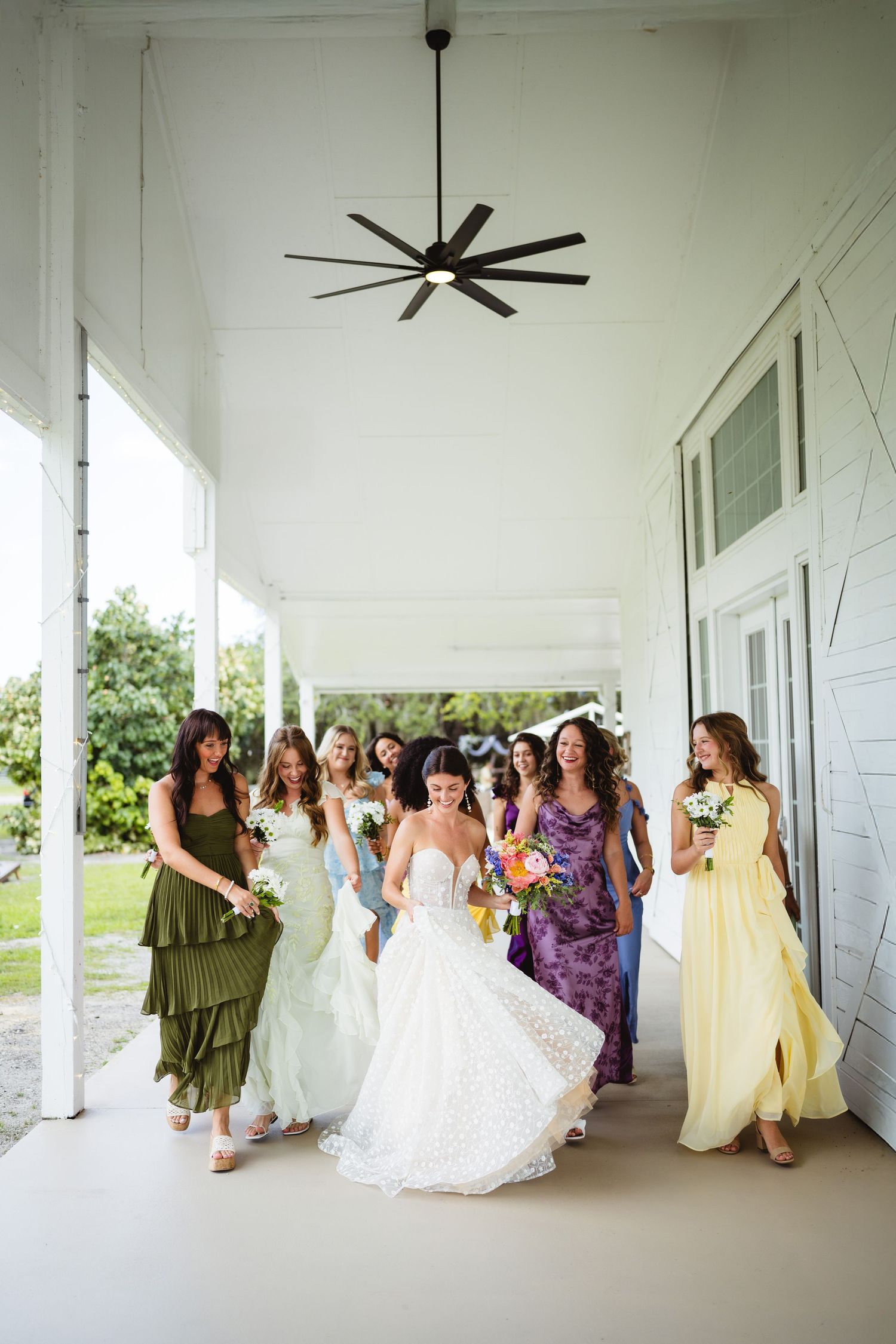 A bridal party wearing colorful dresses walks together on a covered white porch with a ceiling fan above.
