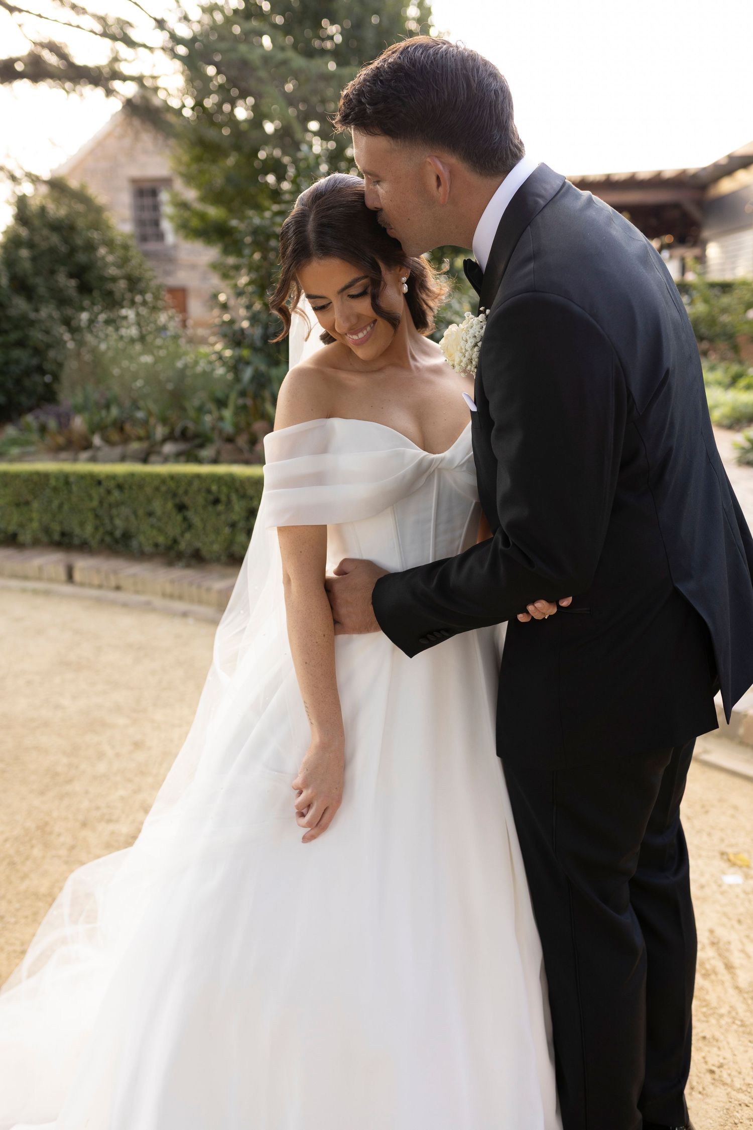 A romantic wedding moment showing a couple embracing outdoors, with the bride in an elegant off-shoulder white gown.