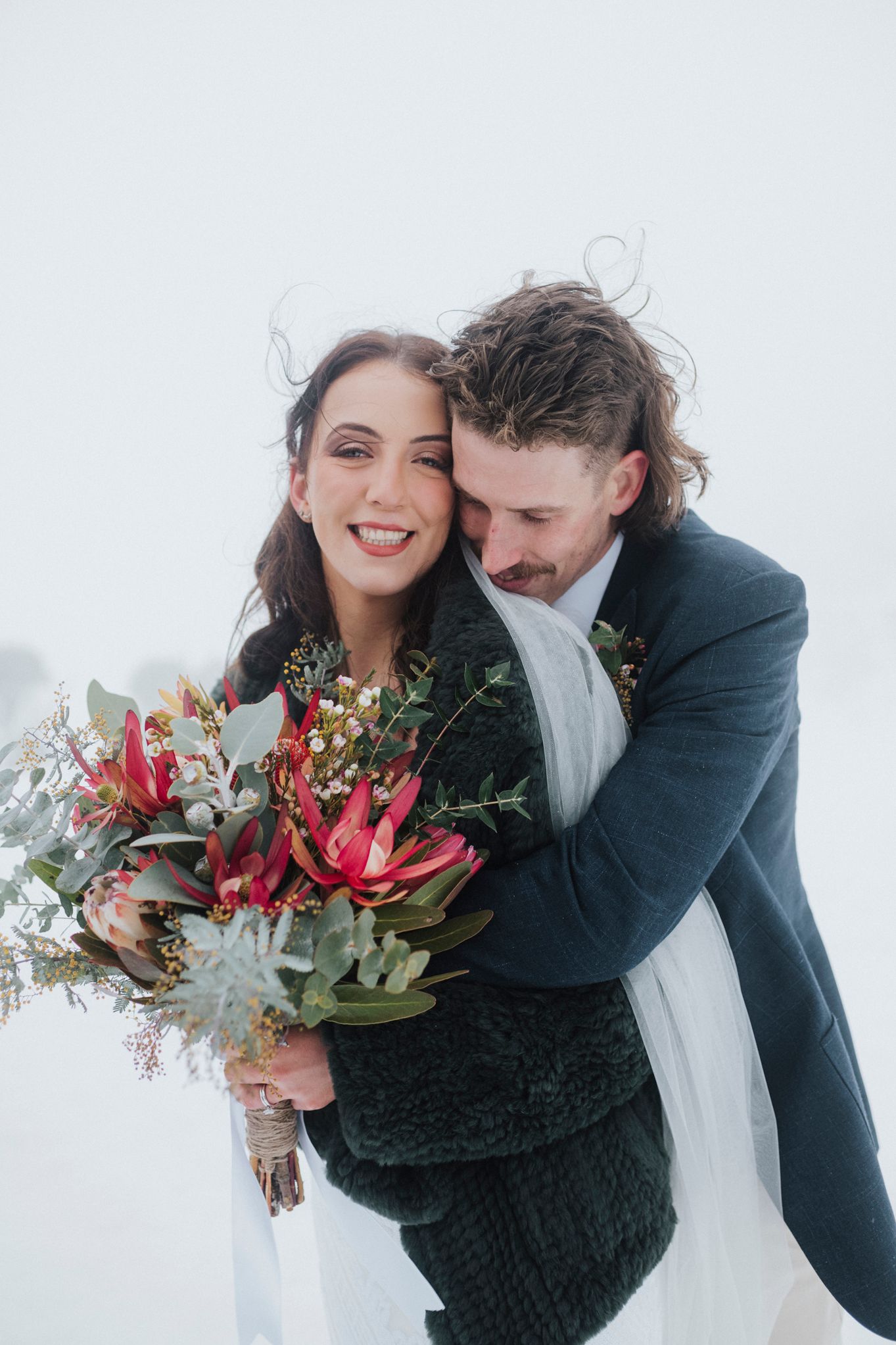 Winter wedding couple with vibrant protea bouquet against white background.