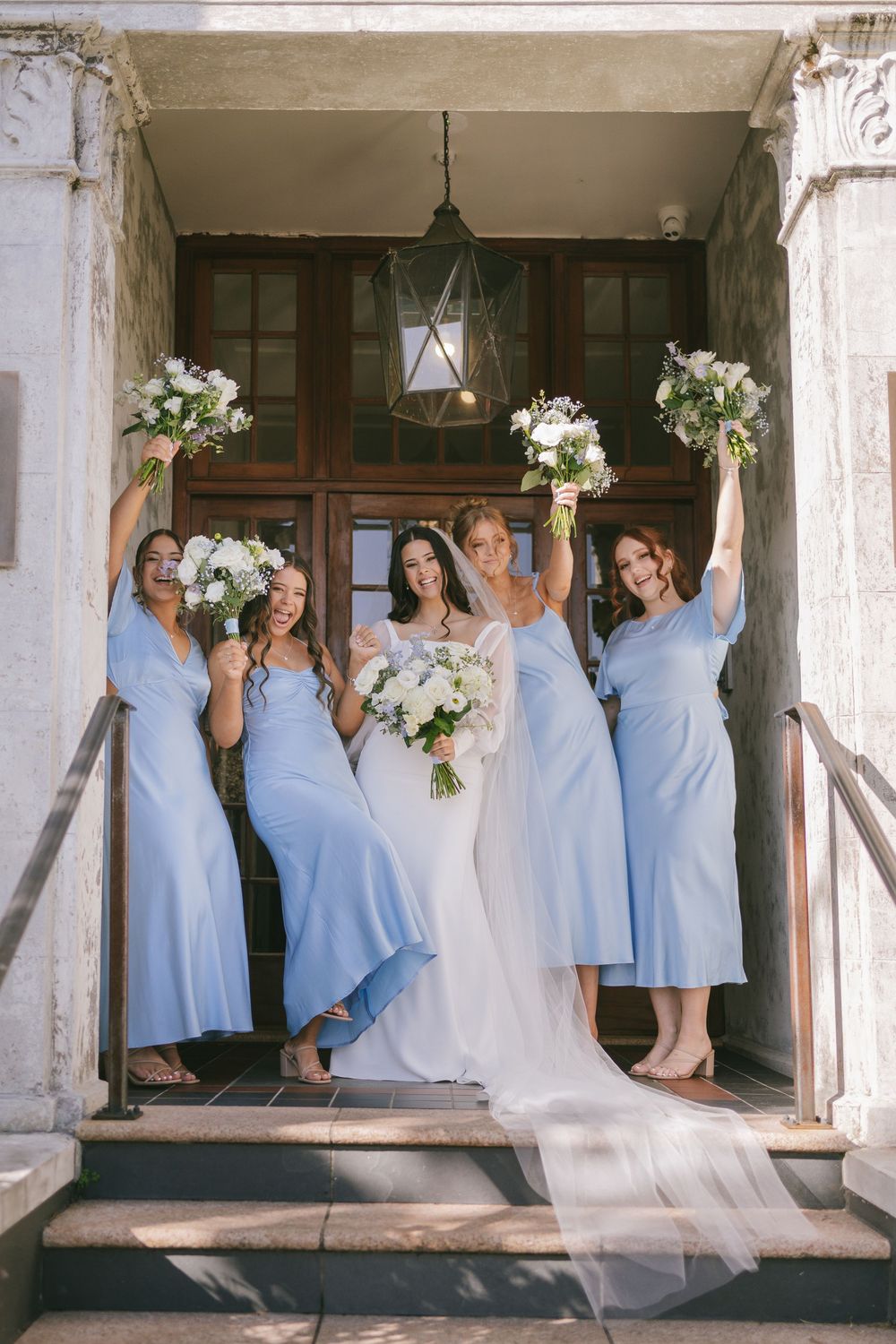 Bridesmaids in light blue dresses pose playfully on a rustic porch.