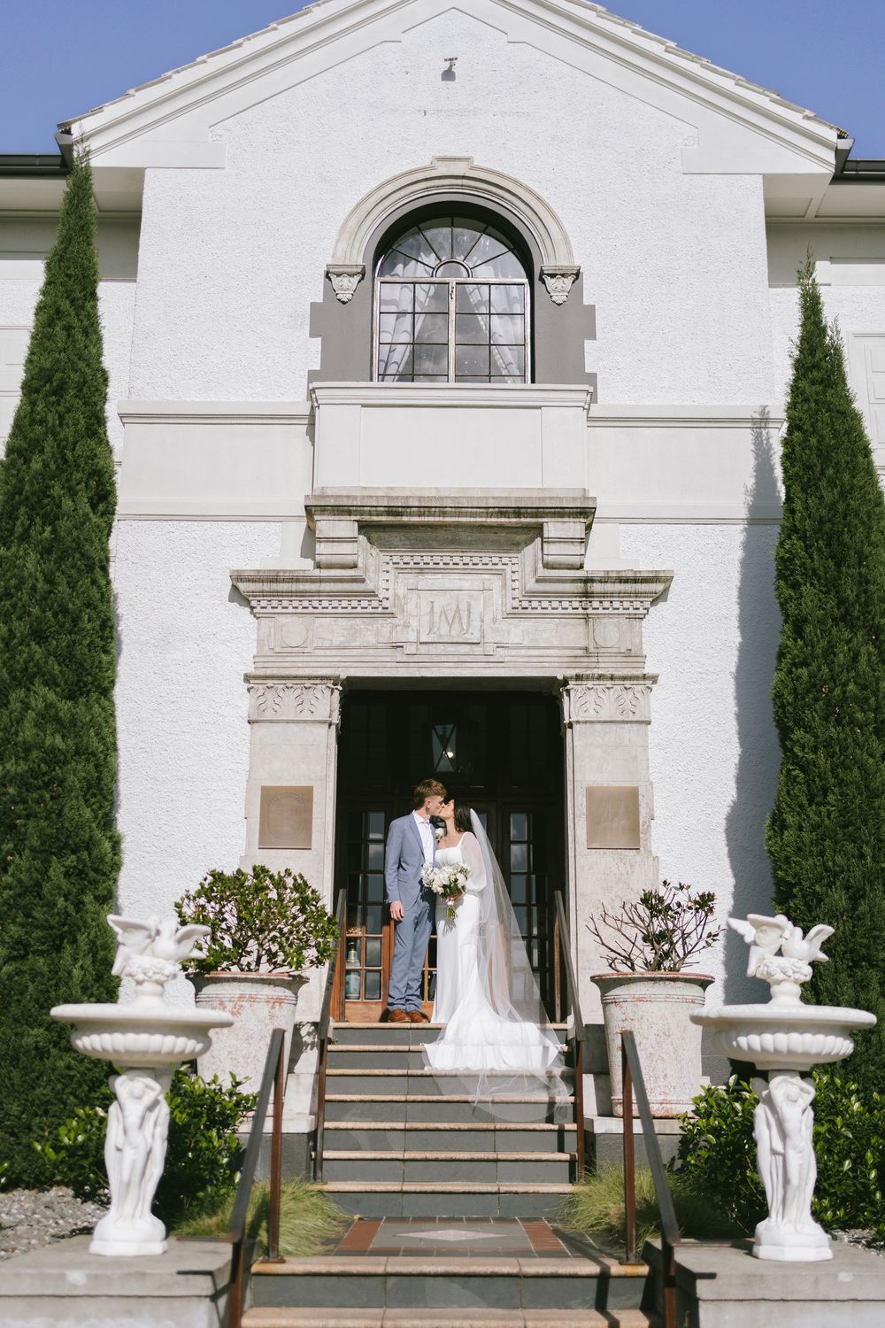 White neoclassical building entrance with cypress trees and decorative statues.