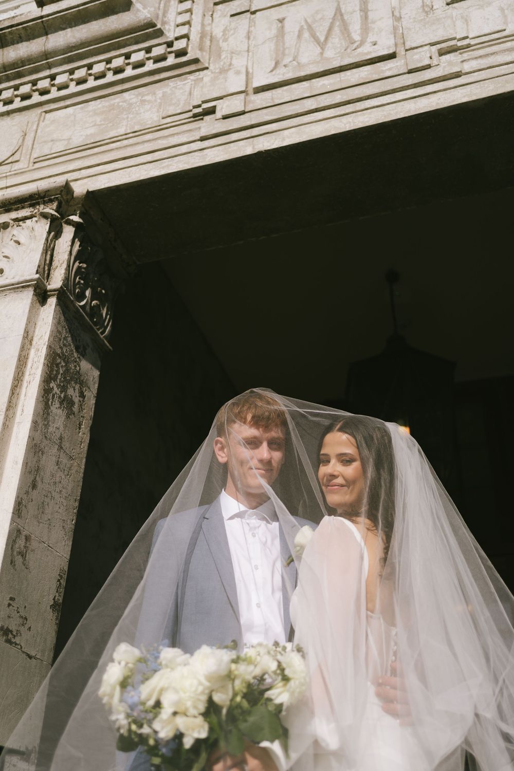 Wedding couple poses under historic stone archway with white floral bouquet.