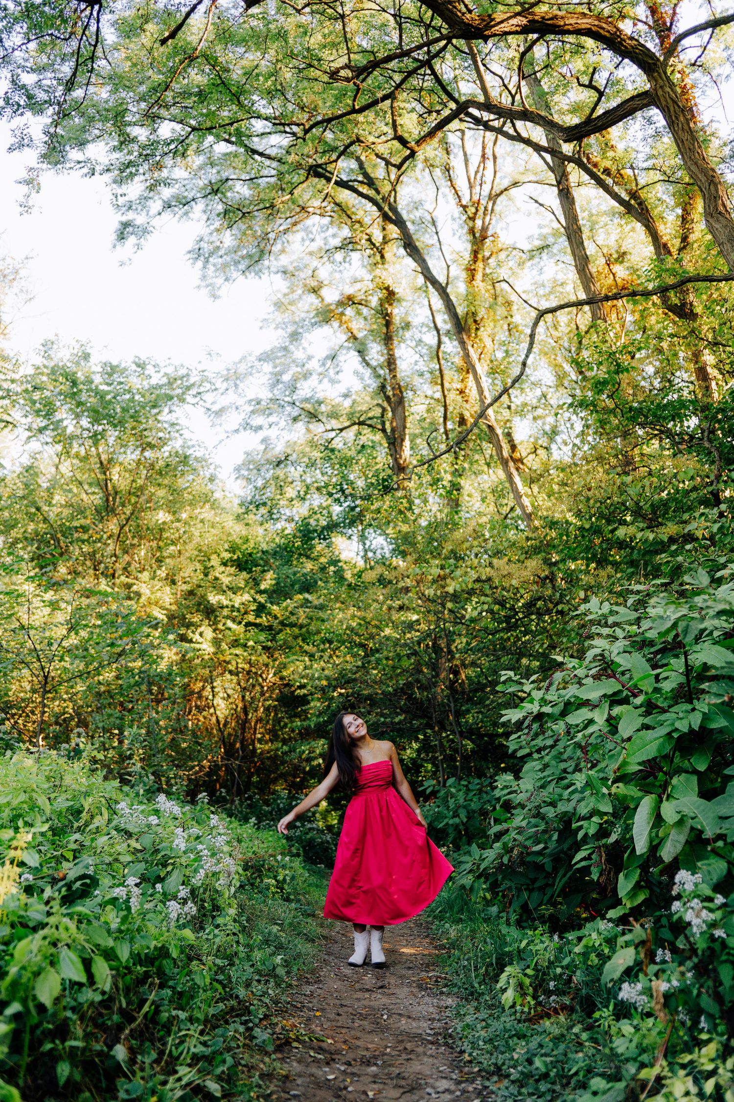 Person in flowing pink dress twirls on forest path surrounded by green foliage.