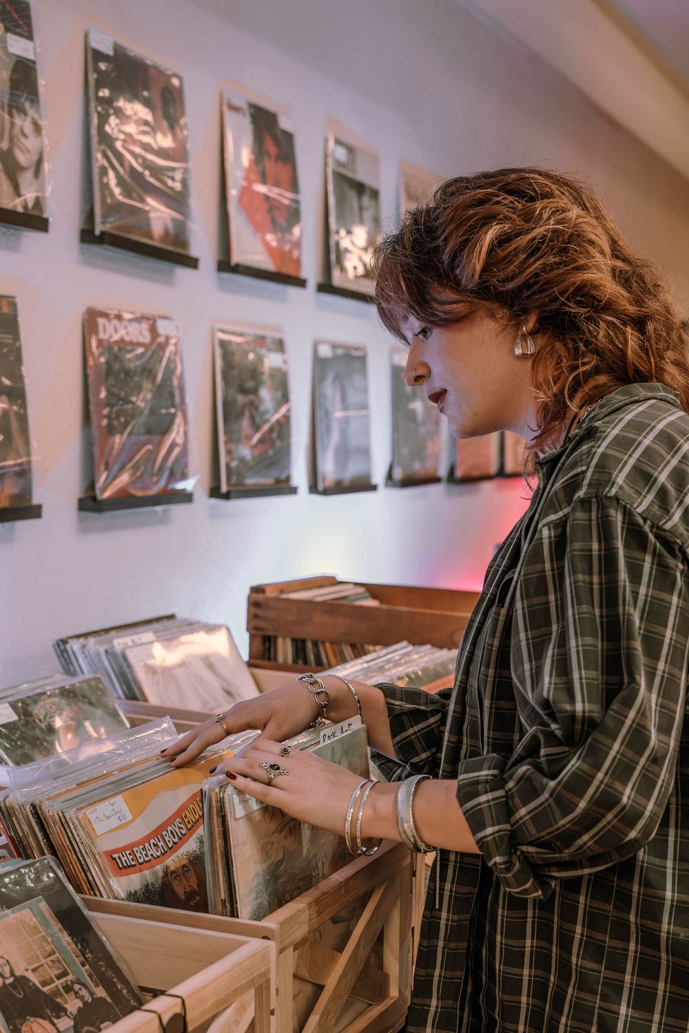 Person in plaid shirt browses through vinyl records at a record store with album artwork displayed on the wall.
