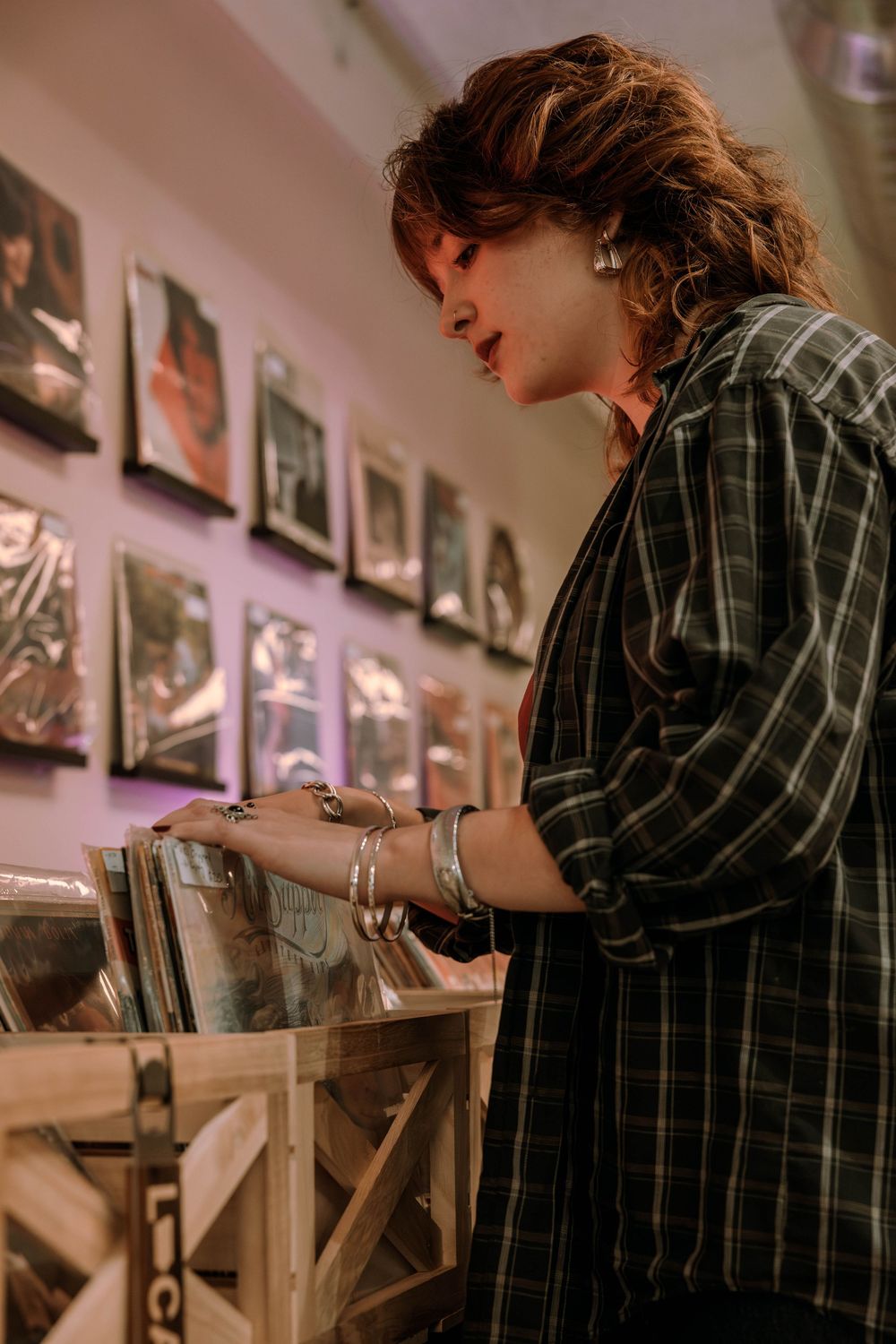 Person in plaid shirt browsing vinyl records at a music store with framed photographs on pink-lit wall.
