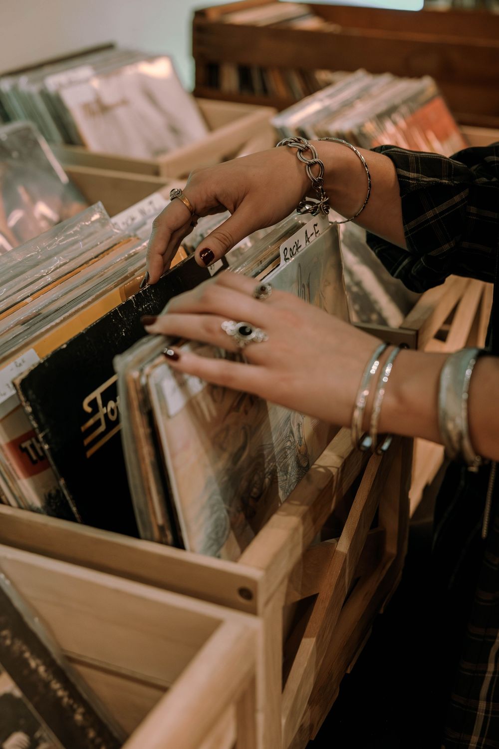 Hands wearing silver bracelets and rings flip through vinyl records in a wooden crate at a record store.