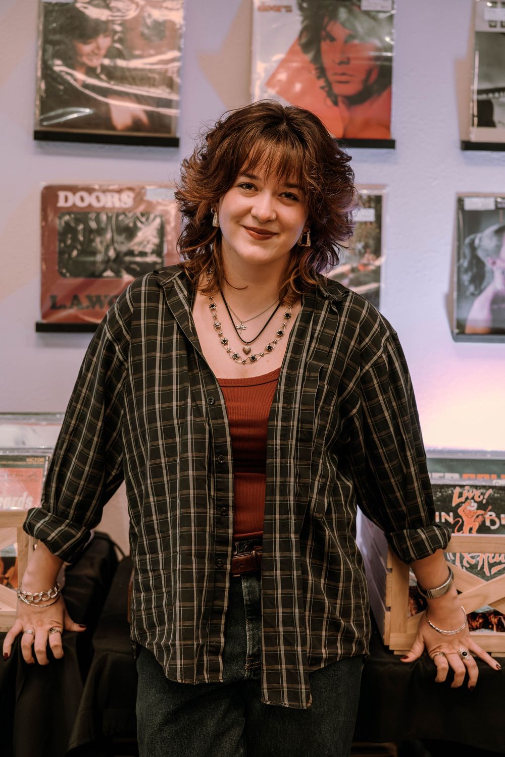 A person in a plaid button-down shirt and layered necklaces poses against a wall with music posters.