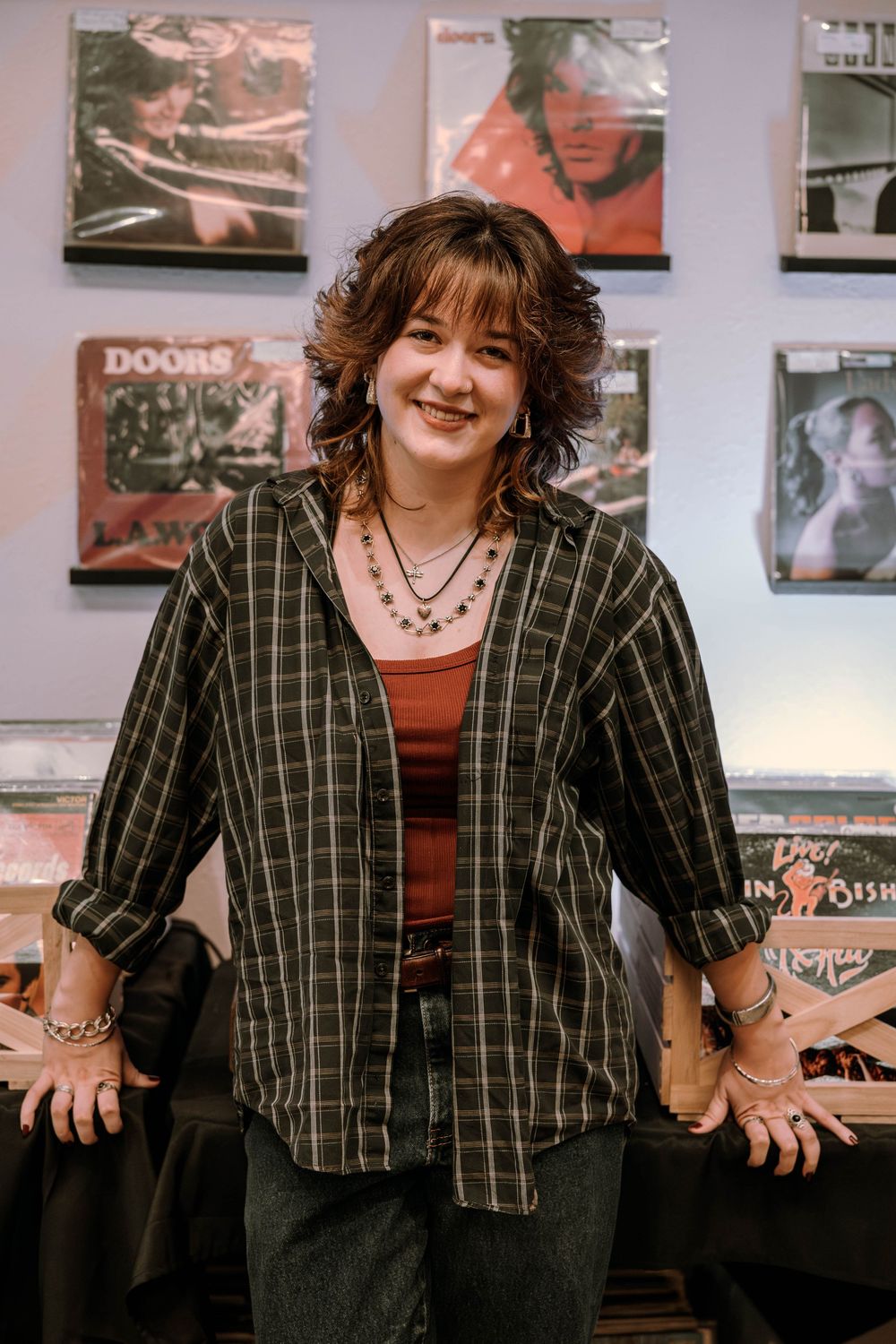 A person in a plaid button-up shirt and layered necklaces stands in front of a wall decorated with vintage album covers.