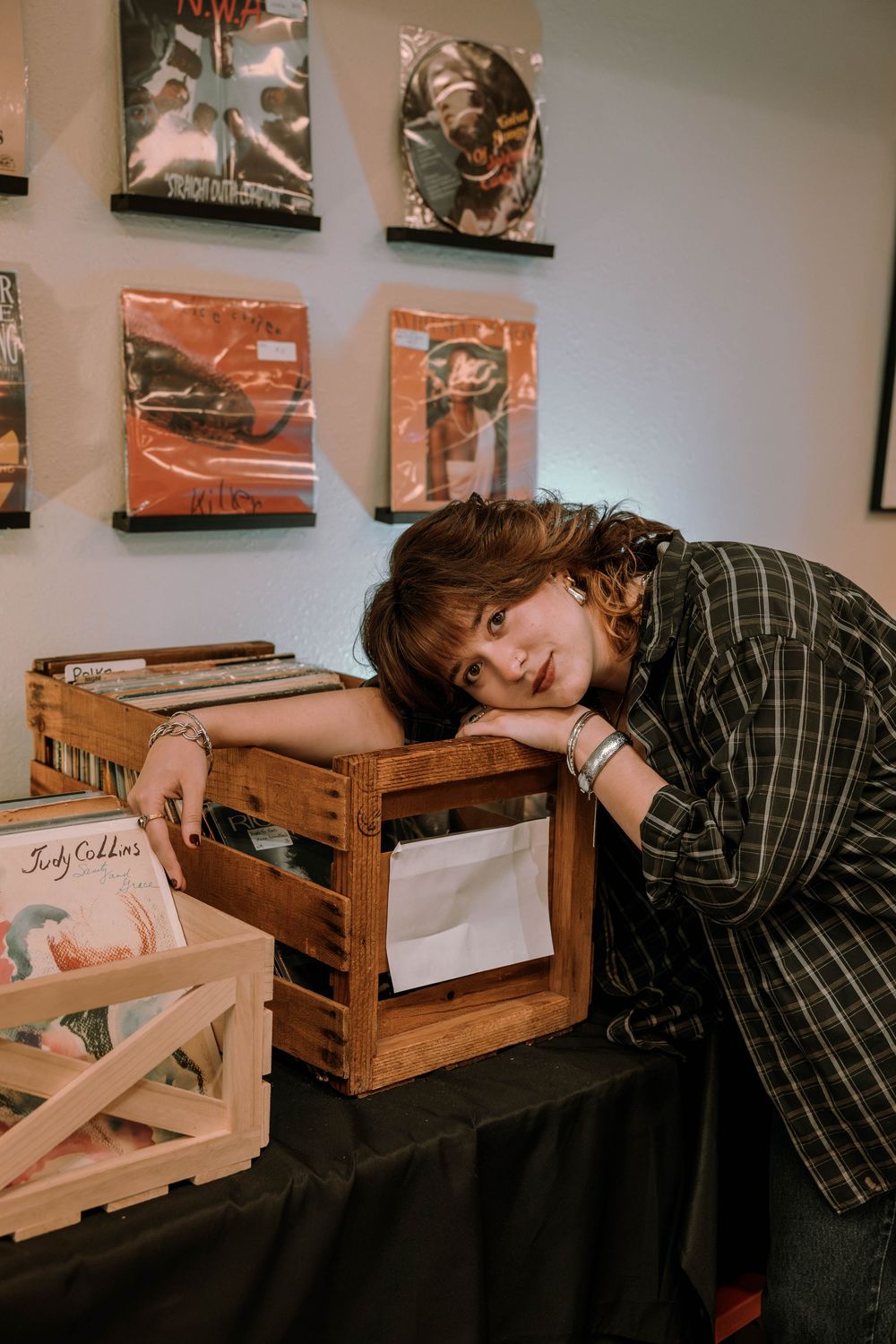 Someone rests their head on a wooden crate filled with vinyl records in a room decorated with album artwork on the walls.