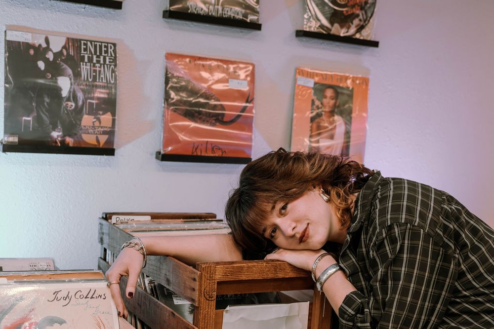 A person in a plaid shirt rests their head on vinyl records with vintage album covers displayed on the pink wall behind them.