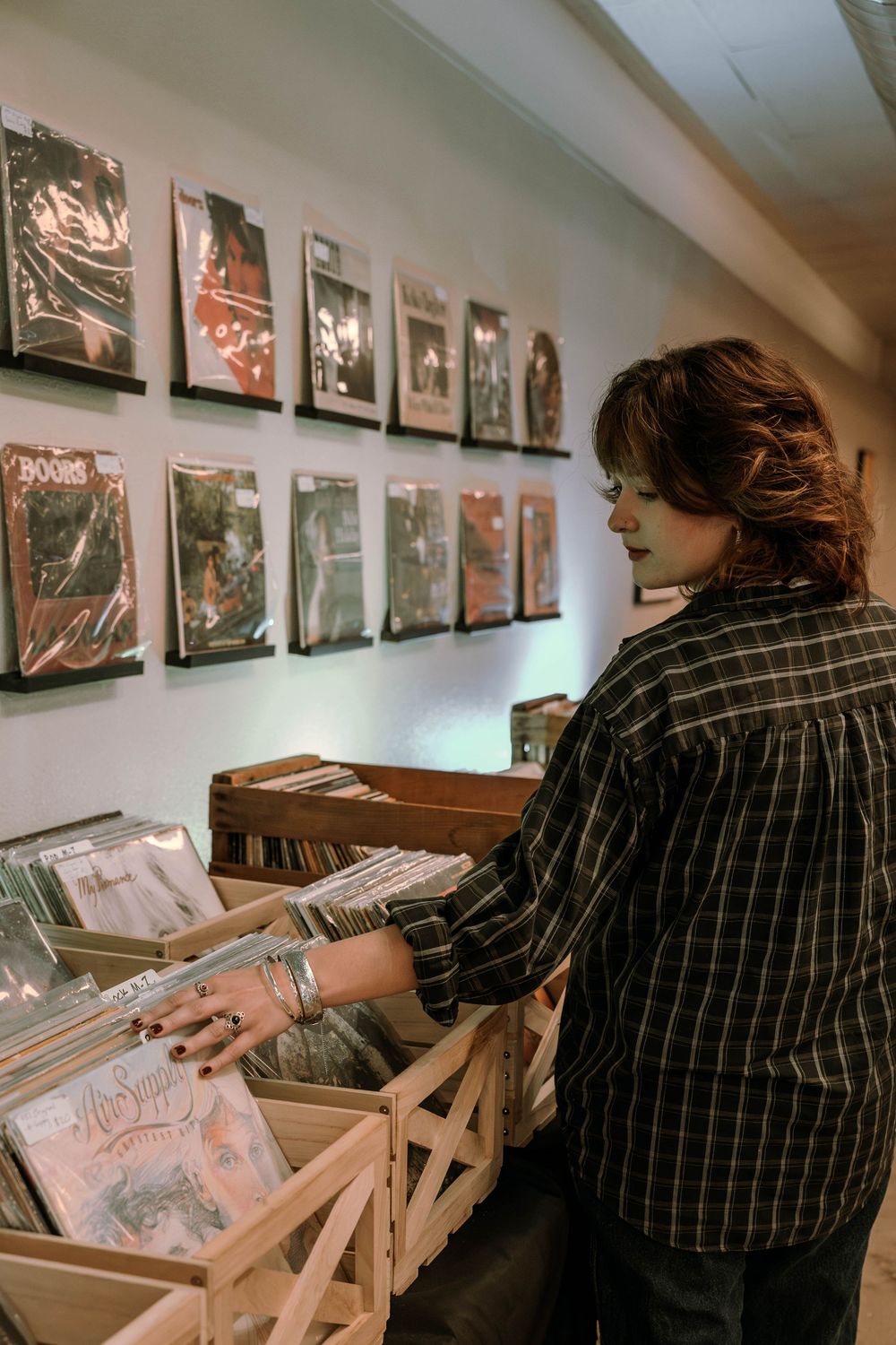 Someone in a striped shirt browses through vinyl records at a music shop with album artwork displayed on the wall.