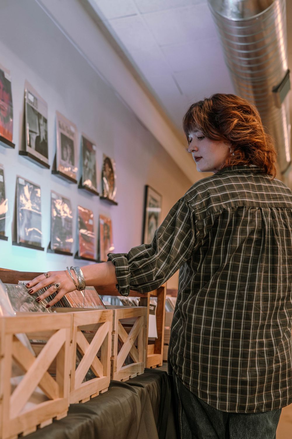 Person in plaid shirt browsing through vinyl records at a shop with framed photos on the wall in background.