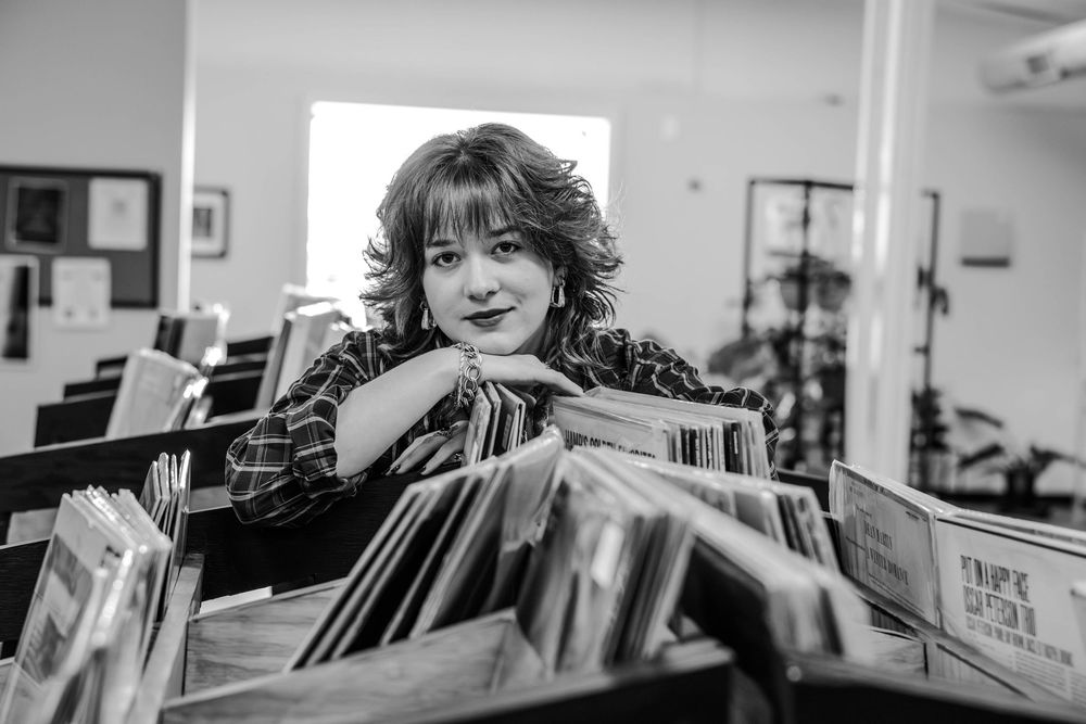 A person flips through vinyl records at a record store with shelves of albums visible in the background.