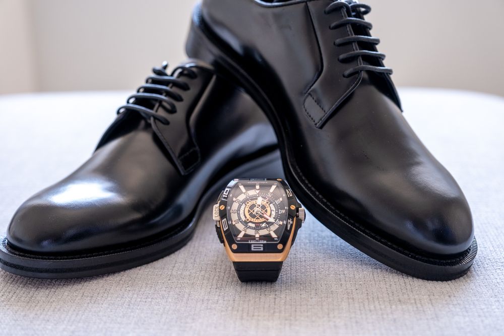 Elegant black leather dress shoes with luxury watch displayed on beige surface.