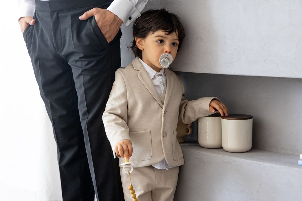 Young child in beige suit standing next to adult legs while holding white container.