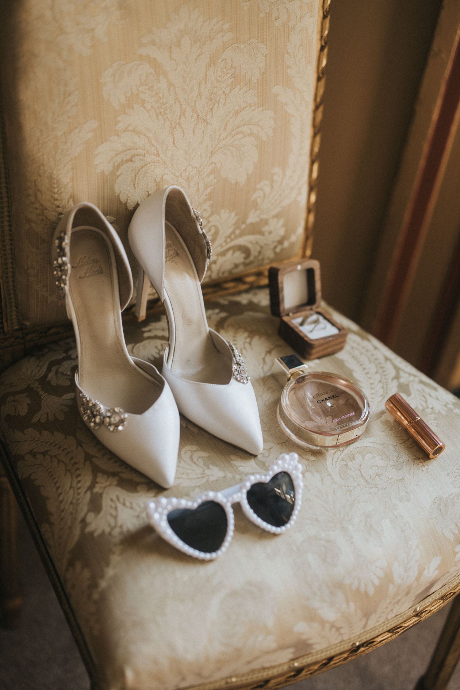 Elegant white bridal shoes with crystal details displayed alongside heart-shaped sunglasses and perfume bottle on damask chair.