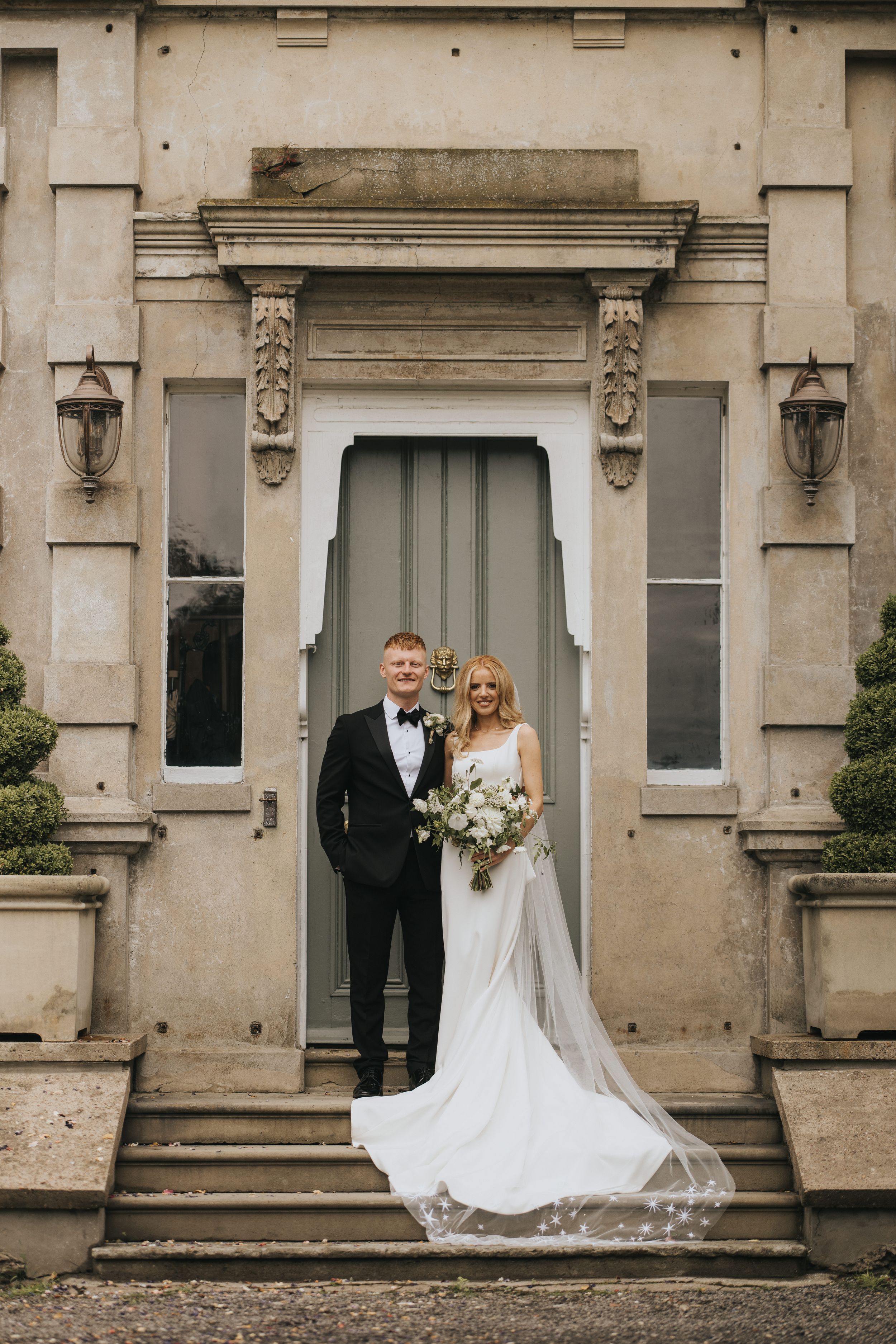 A romantic wedding photo showing a couple posing in front of the elegant stone entrance of Cleatham Hall.