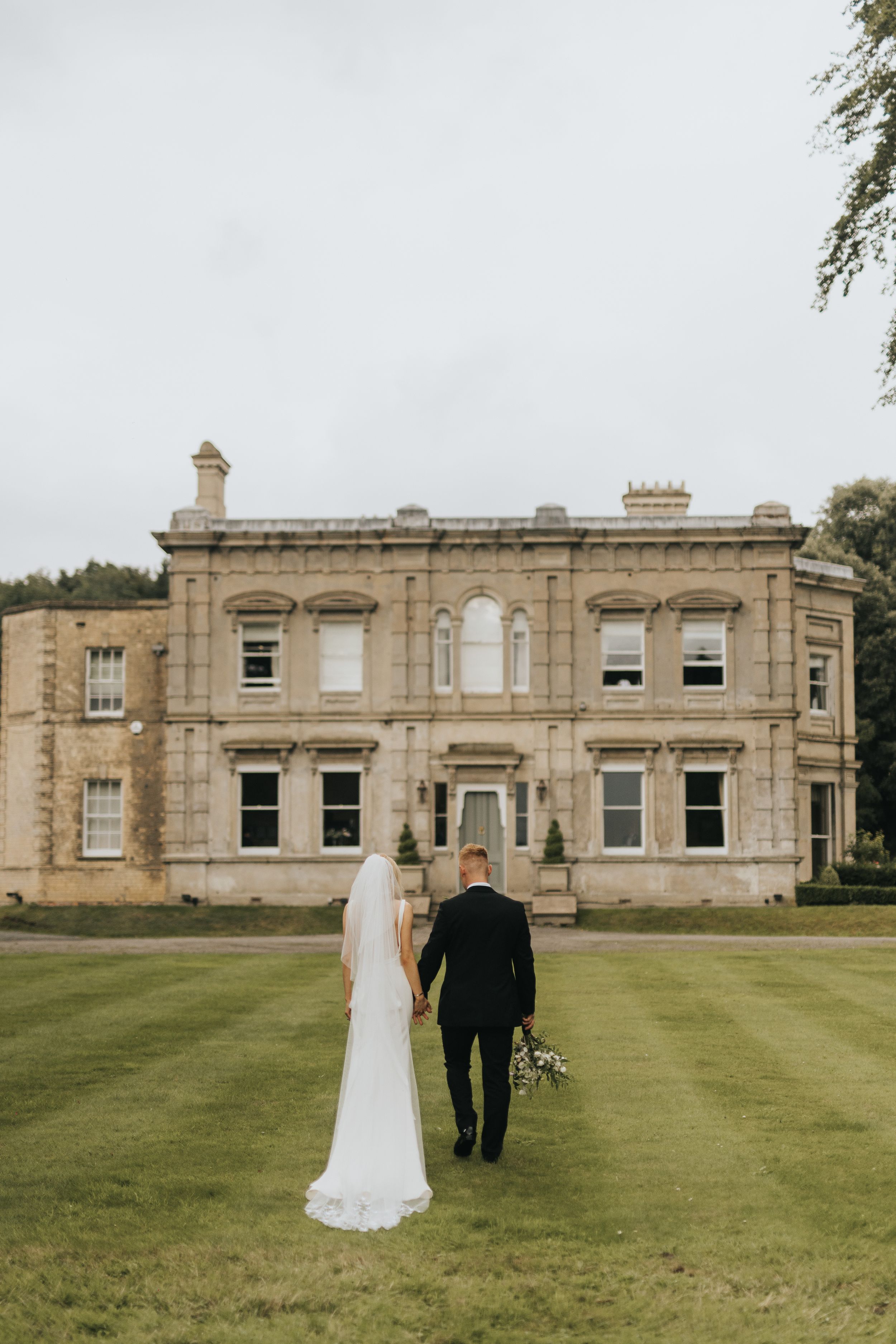 A wedding couple walks across a grand lawn with Cleatham Hall in the background.