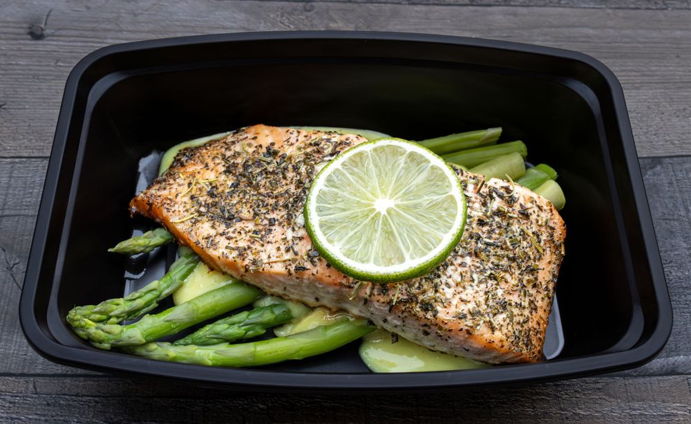 Grilled salmon fillet served in black meal prep containers with asparagus, lime slices, and seasoning.